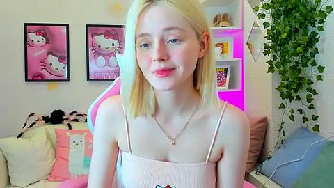 Media: A video of a young, fair-skinned, blonde-haired girl with small breasts, wearing a pink ribbed tank top, sitting in a cozy, pastel-themed room with Hello Kitty decor and plants.