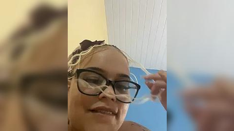 Media: A video of a woman with light skin and a mixed-race appearance, wearing glasses, smiling. She is adjusting a plastic, clear visor. The background features a white ceiling and blue wall.