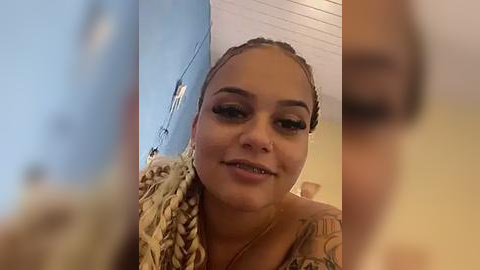 Media: Video of a young, light-skinned woman with braids and tattoos, smiling slightly. She wears a patterned top. Background is blurry, showing a wall and ceiling.