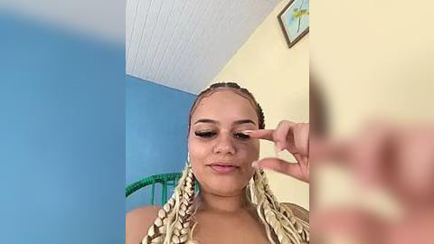 Media: Video of a young woman with light brown skin and braided hair, applying makeup in a room with blue walls and a yellow ceiling.