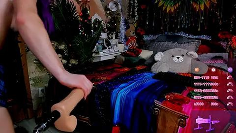 Media: Video of a woman in black high heels, standing beside a plush teddy bear and a red blanket on a bed, in a dimly lit room with festive decor.