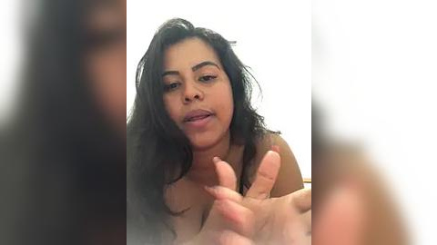 Media: Video of a young woman with long, curly black hair, light brown skin, and full lips, wearing a necklace. She gestures with her hands in a playful manner against a blurred background.