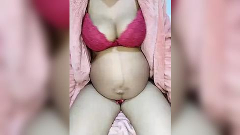 Media: Video of a pregnant woman in a pink bra and robe, showcasing a round belly and light skin tone. Background is blurred.