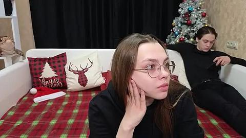 Media: Video of a young woman with glasses, wearing black, resting her head on a couch with a red plaid blanket, next to a man in black pajamas, in a room with a decorated Christmas tree.