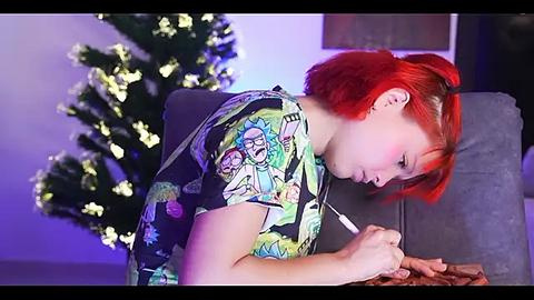 Media: Video of a young woman with red hair, wearing a colorful cartoon T-shirt, drawing on a grey couch, surrounded by a lit Christmas tree and purple ambient lighting.