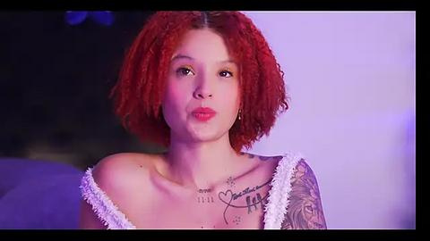 Media: Video of a young woman with light skin and short, curly, fiery red hair. She has a delicate tattoo on her shoulder and wears a white, off-shoulder dress. Background is a blurred, purple-hued gradient.