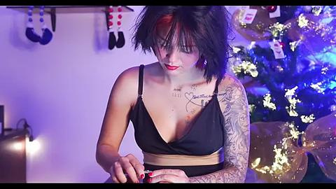 Media: Video of a pale-skinned woman with dark hair, tattoos, wearing a black dress, sitting on a couch near a lit Christmas tree, holding a red object.