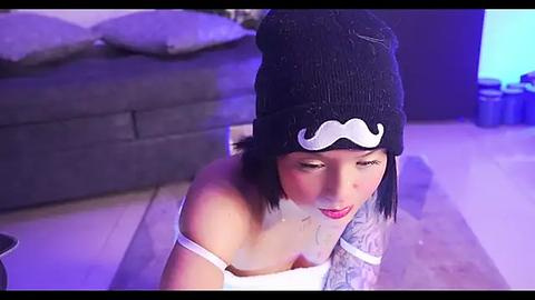 Media: Video of a tattooed woman with pale skin and short black hair, wearing a black beanie with a white mustache, white tank top, kneeling on a gray rug in a dimly lit room with a dark gray sofa and blue furniture.