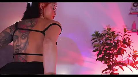 Media: Video of a tattooed woman with long black hair, wearing a revealing black strappy top, sitting in a dimly lit room with pink and purple lighting. She has a large, detailed tattoo on her back.