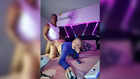 Media: Video of a muscular Black man in a white tank top and beige shorts, standing behind a smiling, slender Asian woman in a blue long-sleeve shirt and beige panties, on a bed with pink sheets in a dimly lit room.