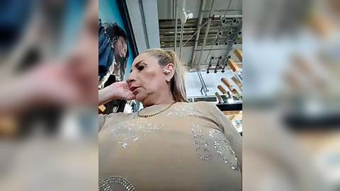 Media: Video of a blonde woman in a beige, sequined dress, with a contemplative expression, leaning on a store window display. Background shows blurred people and store items.