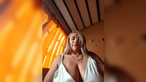 Media: Video of a blonde woman with large breasts in a white, low-cut dress, standing in a dimly lit room with a warm, orange glow and wooden ceiling beams.