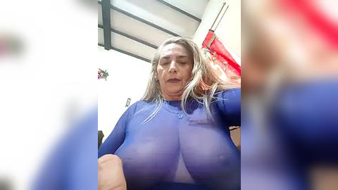 Media: Video of a middle-aged woman with long blonde hair, wearing a tight blue top that accentuates her large breasts, in a cluttered, dimly lit room with a red cloth and metal beams visible in the background.