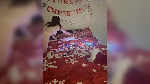 Media: A blurry video of a person in a Santa hat kneeling on a bed with a red Christmas-themed bedspread, illuminated by colorful string lights, in a festive room.