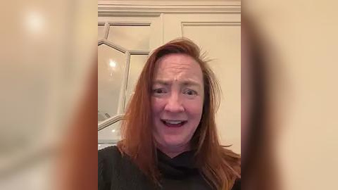 Media: Video of a middle-aged woman with reddish-brown hair, smiling, wearing a black turtleneck, indoors with cream-colored walls and a frosted glass door in the background.