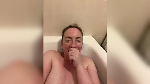 Media: A video shows a naked woman with fair skin, light brown hair, and small breasts, sitting in a white bathtub, holding her chin, with beige tiled walls in the background.