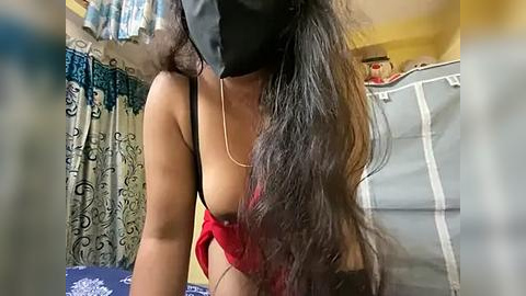 Media: A video of a woman with long, wavy black hair, wearing a black mask, red bra, and a thin gold chain, bending over in a dimly lit room with patterned curtains and a laundry basket.