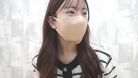 Media: Video of an Asian woman with long brown hair, wearing a beige face mask, black-and-white striped sweater, and a delicate necklace. Background is a semi-transparent curtain with a geometric pattern.