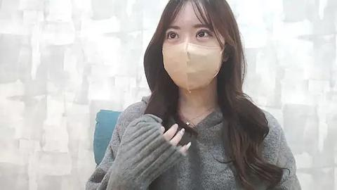 Media: Video of an Asian woman with long, straight brown hair, wearing a gray sweater, a beige face mask, and holding a comb in her right hand, standing in front of a patterned white curtain.