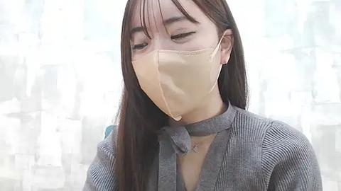 Media: Video of an Asian woman with long brown hair, wearing a gray ribbed sweater, a beige face mask, and a choker necklace, set against a blurred, white, and gray abstract background.