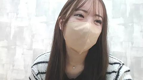 Media: Video of an Asian woman with long brown hair, wearing a beige face mask and black top, against a blurred, abstract white and grey background.