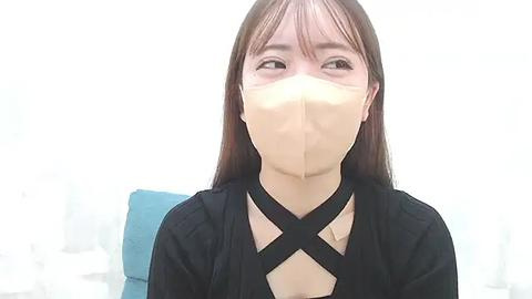 Media: Video of an Asian woman with long brown hair, wearing a beige surgical mask and black crisscrossed cloth mask, sitting in a light blue chair.