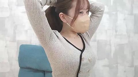 Media: Video of an Asian woman with light skin and brown hair, wearing a beige mask, gray knit sweater, and blue chair in a room with white, abstract-patterned curtains.