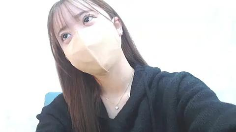 Media: Video of a young Asian woman with straight, dark brown hair and light skin, wearing a black mask covering her nose and mouth, and a black sweater. Background is plain white.