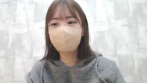 Media: Video of an East Asian woman with shoulder-length dark brown hair, wearing a beige face mask and a grey hoodie, standing against a white and grey abstract-patterned backdrop.