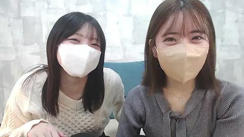 Media: Video of two Asian women with light skin, straight black hair, and beige masks, wearing patterned tops, seated against a white and blue patterned background.