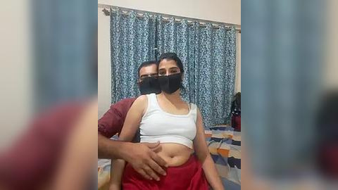 Media: Video of a South Asian couple in a bedroom, man behind woman wearing red shirt, woman in white crop top, black mask, blue curtains, bed with blue and white patterned sheet.