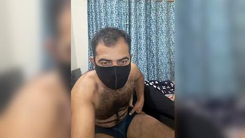 Media: Video of a shirtless South Asian man with short hair, wearing a black face mask and dark shorts, sitting on a bed in a room with blue patterned curtains.