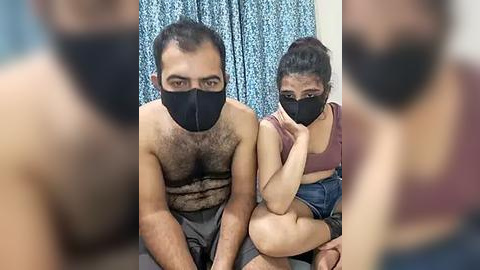 Media: Video of a man with hairy chest and a woman with dark hair, both wearing black face masks, sitting closely in a room with blue-patterned curtains.