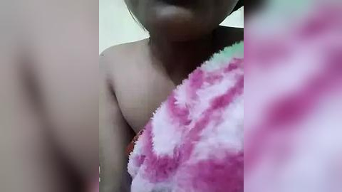 Media: Video of a young person with light brown skin, partially covered by a pink and white fluffy towel, focusing on their upper body and face, with blurred background.