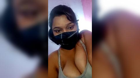 Media: A video of a woman with dark skin and long black hair, wearing a black face mask and a revealing gray tank top, with her cleavage partially visible. The background is blurred, focusing on her face.