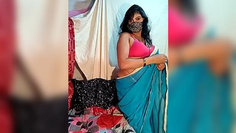 Media: A video of a woman in a pink bra, black mask, and blue sari, sitting on a colorful quilt, indoors, blurry background.