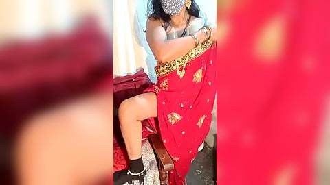 Media: Video of a woman in a red saree, with a leopard print mask, sitting on a bed, legs crossed, in a dimly lit room.