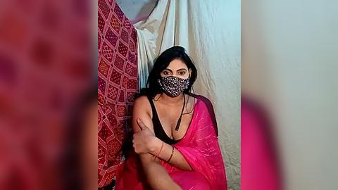 Media: Video of a young woman with medium skin tone, wearing a pink saree, black mask, and black bra, sitting against a tent wall, with blurred red and pink fabrics in the foreground.