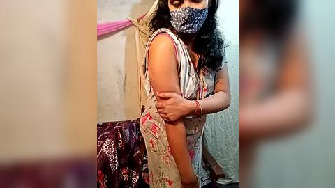 Media: Video of a woman in traditional Indian attire with a floral saree, mask, and jewelry, adjusting her saree in a dimly lit room with beige walls and a pink towel.