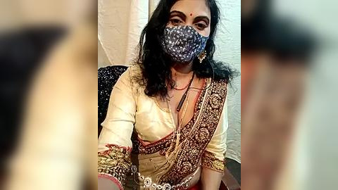 Media: A video of an Indian woman in a traditional saree, with a patterned mask covering her face, sitting on a chair. The background features a white wall and blurred people.