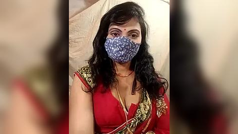 Media: Video of a South Asian woman in a red sari with gold embroidery, wearing a blue patterned face mask, sitting indoors against a beige wall.