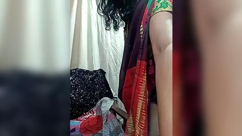 Media: Video showing a partially obscured person in a red and gold saree with a white blouse, standing against a white curtain. The background includes a dark, speckled bag and a partially visible person in the foreground.