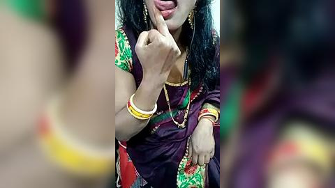 Media: Video of a young woman in a purple sari with green accents, wearing gold jewelry, including a necklace and bangles, playfully sticking her tongue out and covering her mouth with her finger.