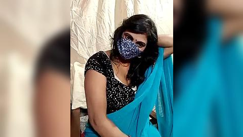 Media: Video of a South Asian woman with medium skin tone, wearing a blue saree and black mask, sitting on a white textured cloth. Background shows a blurred figure in a blue shirt.