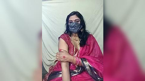 Media: Video of a South Asian woman in a red saree, wearing a black face mask and gold jewelry, sitting against a beige backdrop.