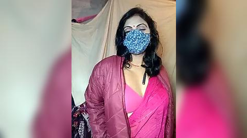 Media: Video of a woman with fair skin, wearing a blue face mask, pink jacket, and a low-cut pink top, standing in front of a beige backdrop, blurred background.