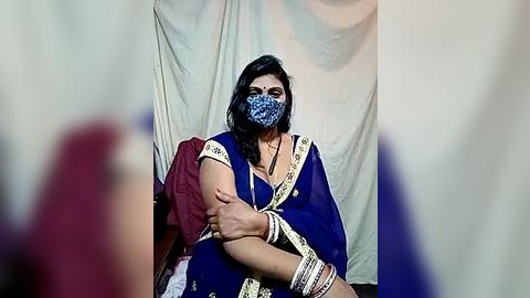 Media: Video of a South Asian woman with medium brown skin, wearing a blue sari, patterned mask, and gold jewelry, seated in a simple room with a white curtain backdrop.