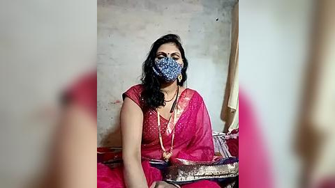 Media: Video of a woman in a bright pink sari with a blue patterned face mask, seated on a bed with a red sheet, wearing a gold necklace, in a simple room with a beige wall and white curtain.