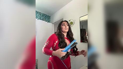 Media: A video of a woman in a red, tight-fitting bodysuit with long, wavy brown hair, holding a phone in a modern, brightly lit bathroom with white walls and blue-patterned wallpaper.