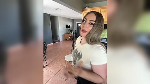 Media: A video of a light-skinned woman with shoulder-length, straight, light brown hair, wearing a white graphic t-shirt and black pants, sitting in a modern kitchen with wooden cabinets, a tiled floor, and a microwave.